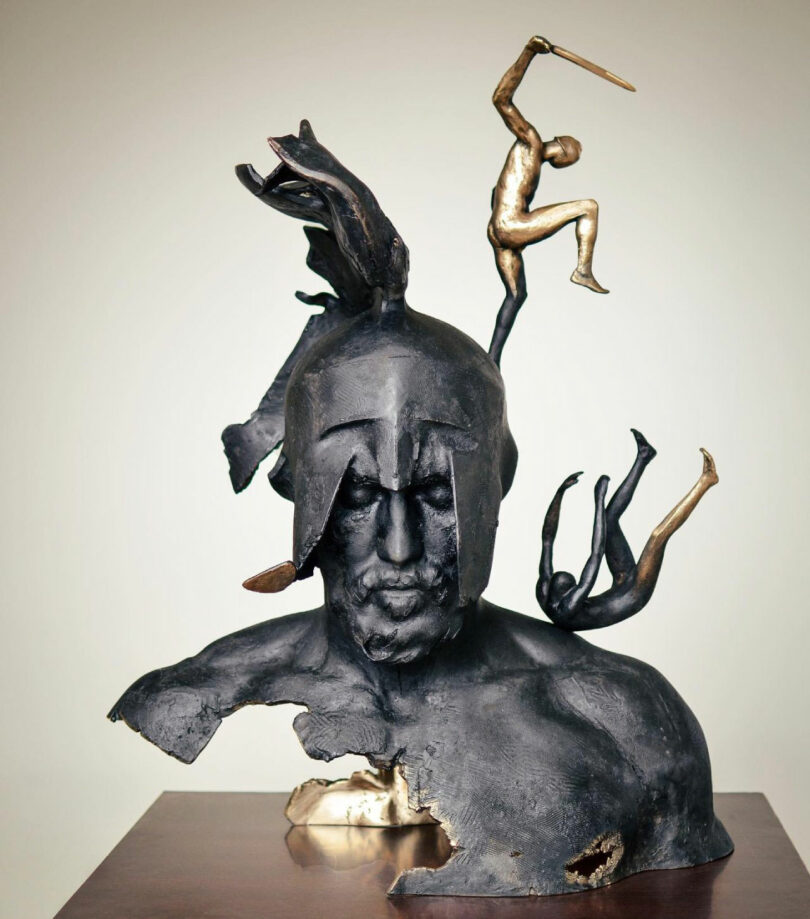 A bronze sculpture by Neil Ferrier depicts a helmeted male bust with abstract, fragmented features and small human figures interacting dynamically atop and around the head.