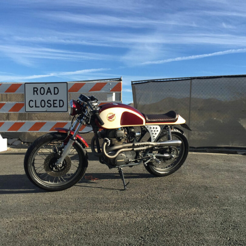 A vintage motorcycle, once owned by Neil Ferrier, is parked on asphalt in front of a "Road Closed" sign and a barricade, with a chain-link fence and mountains in the background.