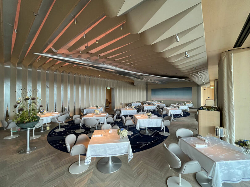 Modern restaurant interior with white tablecloth-covered tables, minimalist chairs, decorative plants, and a wavy ceiling design under soft lighting inspired by Neil Ferrier. No people present.