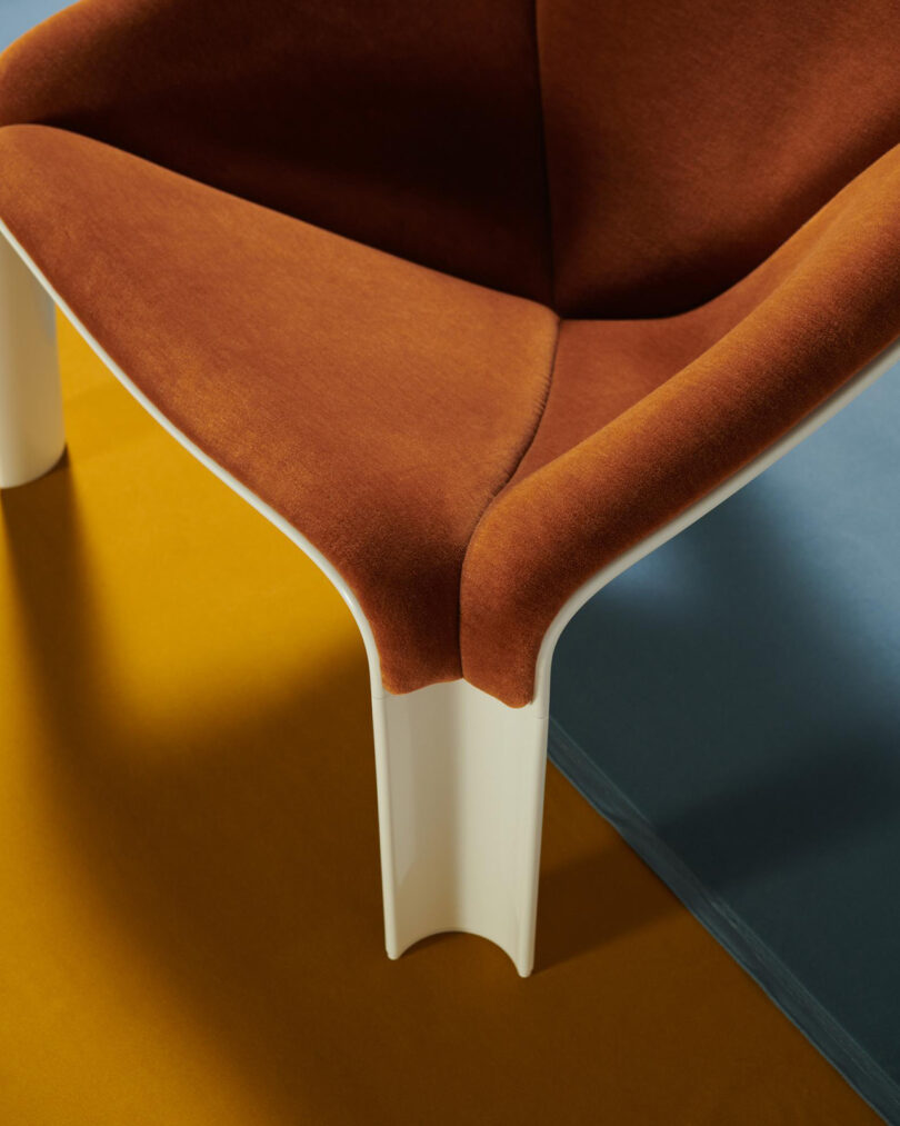 A close-up view of a modern chair with rust-colored upholstery and white legs, placed on a surface divided into mustard yellow and blue sections.