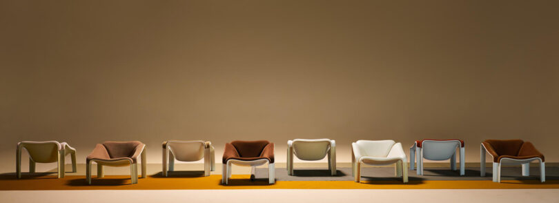 A row of eight modern lounge chairs in various earth tones and white, aligned in front of a plain beige wall on a color-blocked floor.