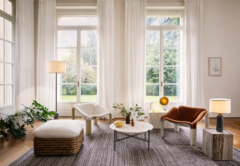 A modern living room with large windows, white curtains, three armchairs, a marble coffee table, plants, and table lamps on a textured rug.