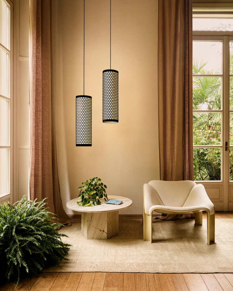 A modern lounge area with a low chair, round marble coffee table with a plant and book, two pendant lights, and large windows draped with sheer curtains.