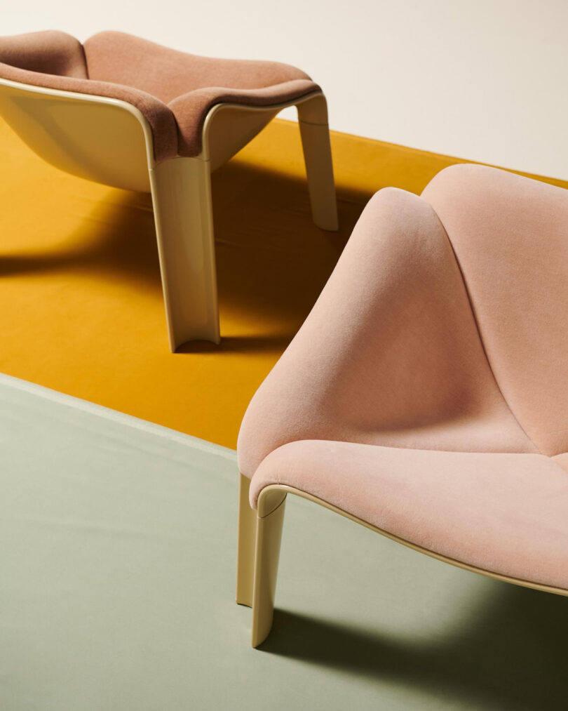 Two modern, sculpted chairs with pale pink upholstery and cream-colored legs are positioned on a mustard yellow and light green surface.