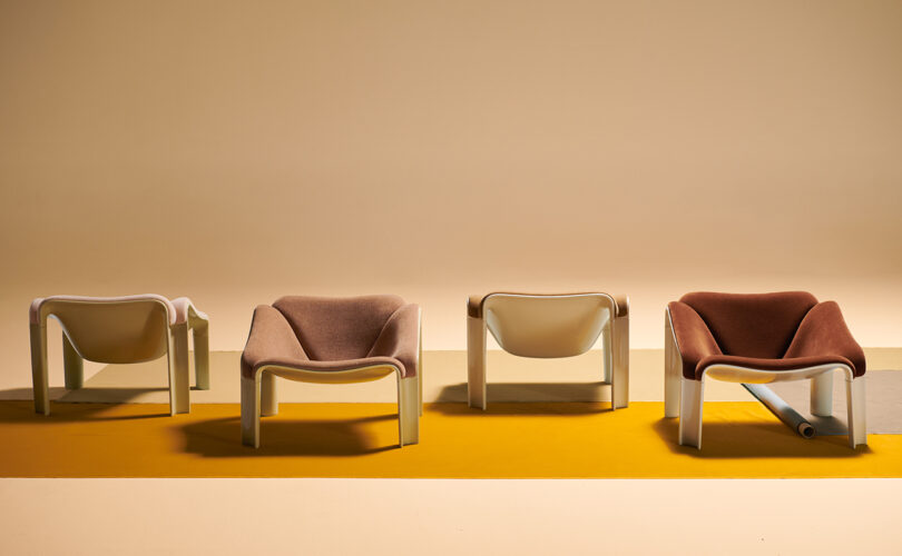 Four modern lounge chairs with white frames and brown or pink upholstery are arranged in a row on a yellow and beige floor against a beige background.