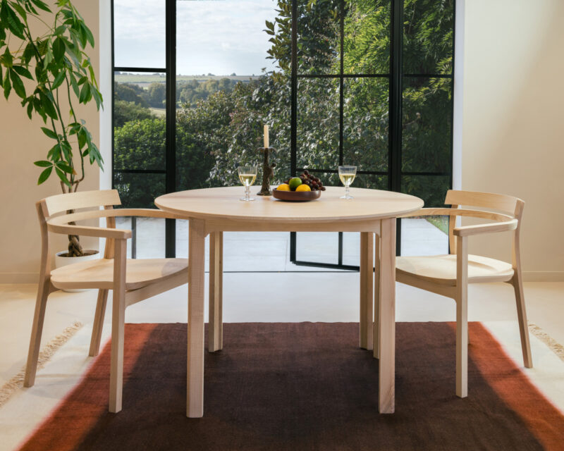 Light wood round dining table with matching armchairs, set on a gradient rug in front of large windows overlooking greenery.