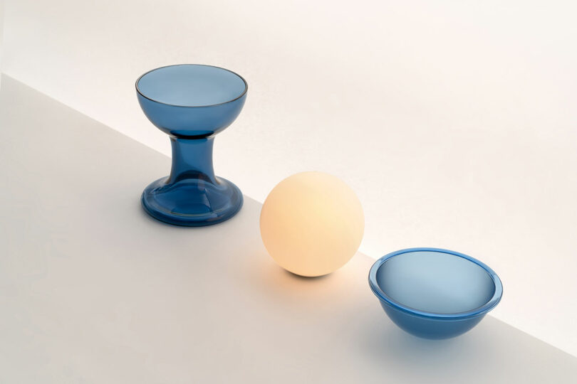 A blue goblet, a glowing Gelato Lamp by Carlo Nason, and a blue bowl are arranged on a white and beige surface.