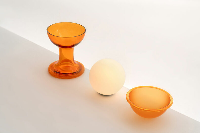 An orange glass goblet, a white spherical object reminiscent of a Gelato Lamp by Carlo Nason, and an orange glass bowl are arranged on a white surface with a diagonal shadow.