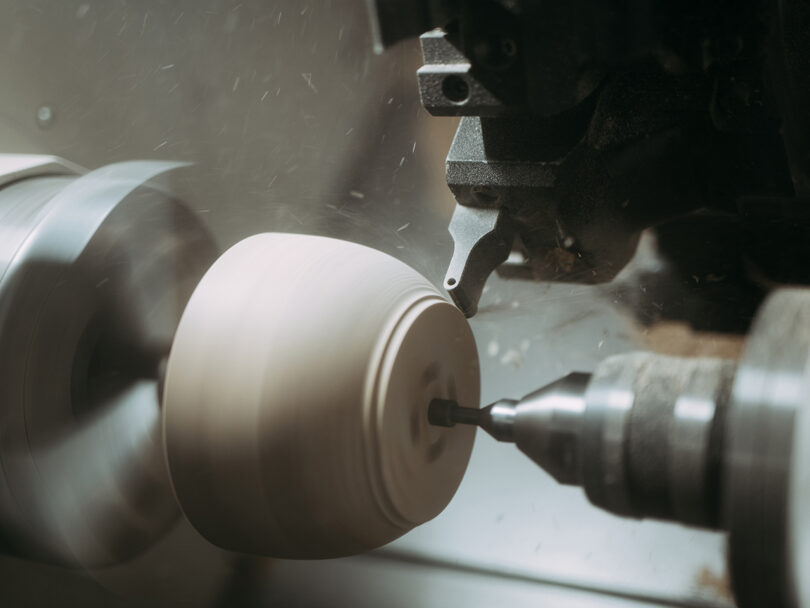 Close-up of a wood lathe machine shaping a spinning cylindrical piece of wood, with wood shavings and dust visible in the air.
