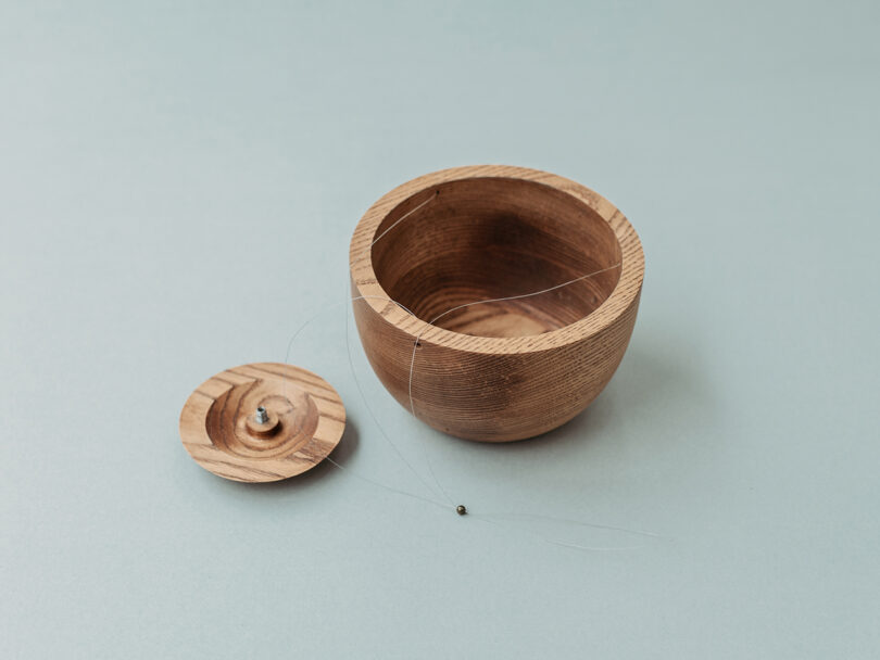 A round wooden container with its lid removed, showing a fine fishing line and a small metal sinker attached inside, set on a light blue background.