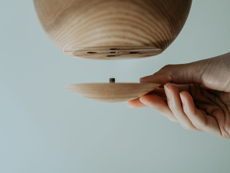 A hand holds a wooden disc with a screw, aligning it with the bottom of a larger wooden object against a plain background.
