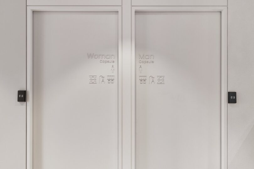 Two adjacent white labeling doors "The capsule woman" and "Man's capsule," with symbols below and key plates next to the doors of the far left and far right