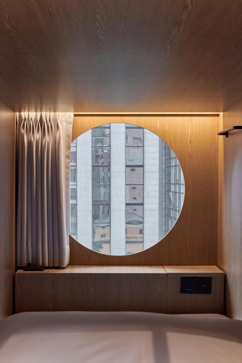 A round window on the other end of the capsule pod offers a view of the city building, placed in a wooden wall above a bed; White curtains are pulled to one side and electrical outlets are visible under the right window