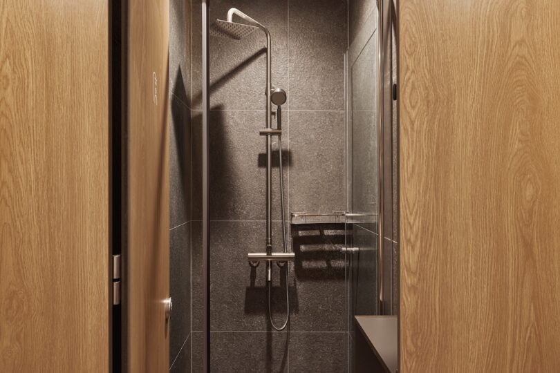 A modern hotel shower with a shower head and a glass door, surrounded by dark tiles and light wooden doors