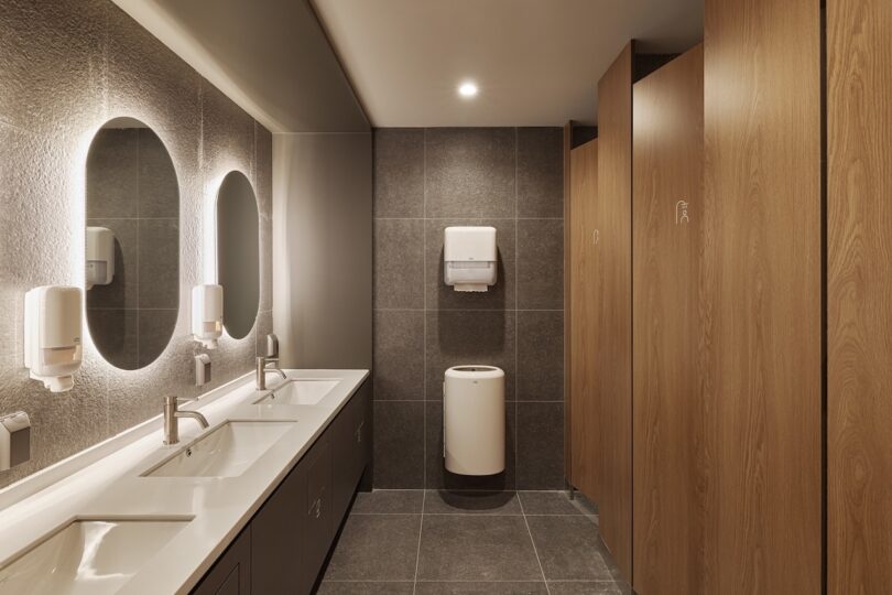Modern Public Public Restroom for Guests with Capsule Reservations at Hotel Drip & Drop Featuring Three Wooden on the Right, Two Sinks with Mirrors Above and Wall-Mounted Soap Dispensers, Paper Towel Distin and Trash Can Mounted On The Back Back Middle Wall, and gray tiled floors and walls