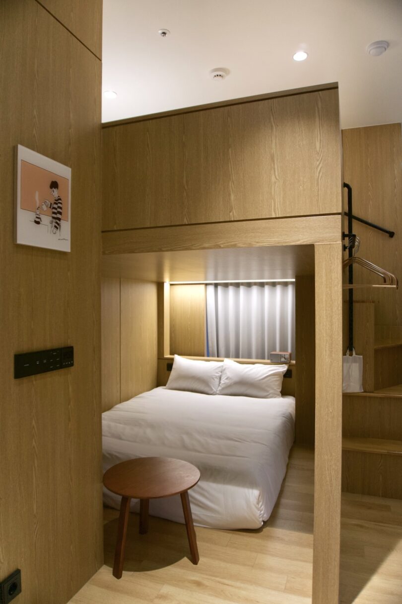 A modern, compact bedroom with wooden walls, a white double bed, a small round table, a loft bed above, and hangers on an open rack. A picture of a cartoon man pouring coffee hangs on the wall to the left.