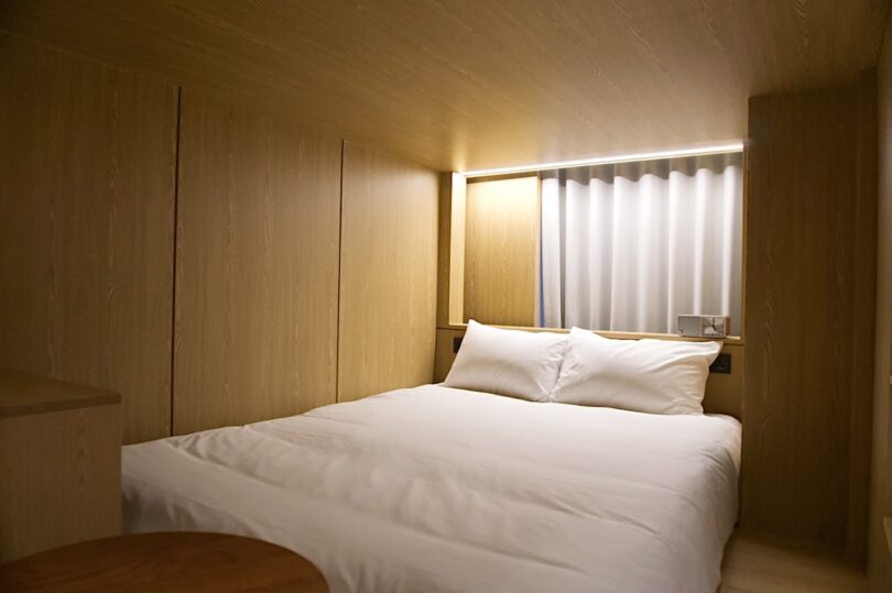 A modern bedroom at HOTEL DRIP&DROP features wood-paneled walls, a double bed with white bedding, and a window with vertical blinds above the headboard