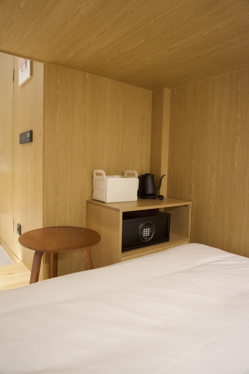 A small wooden room featuring a bed, a round stool, a shelf with a white container, a black electric kettle, and a safe at the foot of the bed