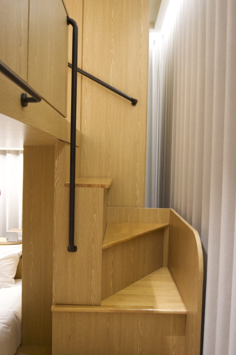 A narrow wooden staircase with black handrails leads to an upper bunk in a compact HOTEL DRIP&DROP room with light wood paneling and floor-to-ceiling curtains