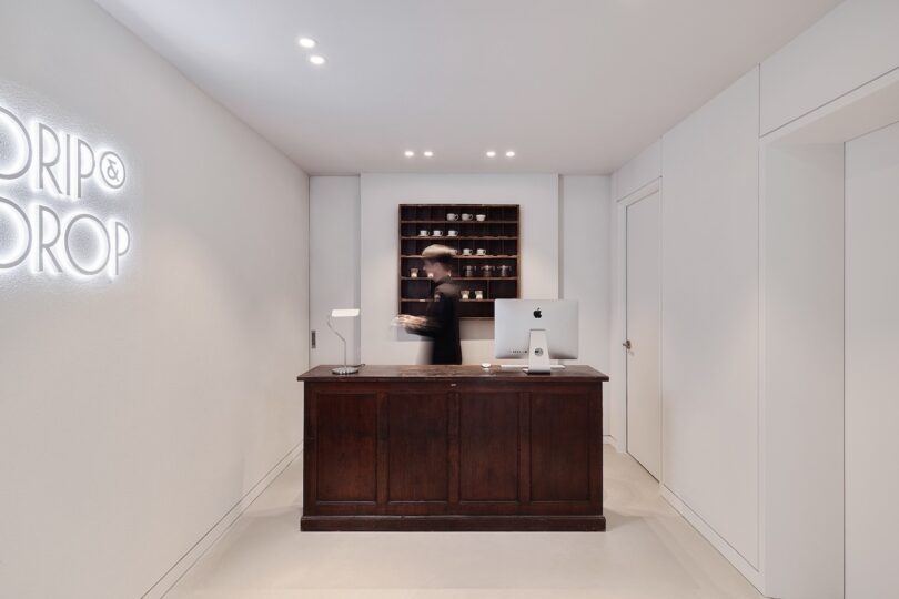 A person stands behind the dark wooden reception desk at the Drip & Drop hotel, with a computer, shelves and coffee beans in the back containers and "Drip & drop" illuminated on the white wall on the left