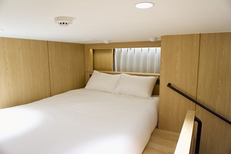 A neatly made bed with white linens in a small, wood-paneled room in the loft at HOTEL DRIP&DROP, featuring a low ceiling and minimal lighting