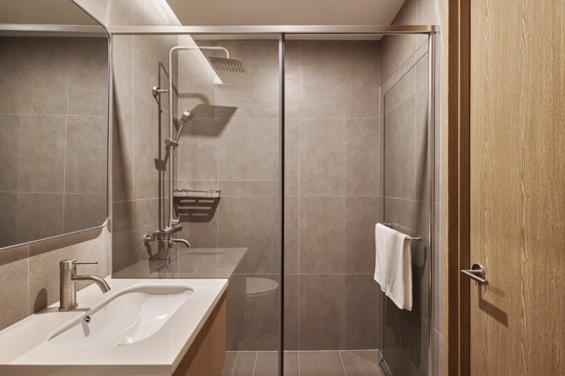 The modern hotel bath has a closed glass shower, a shower with precipitation mounted on the wall, gray tiles, rectangular sink, large mirror and a towel hanging on a rail