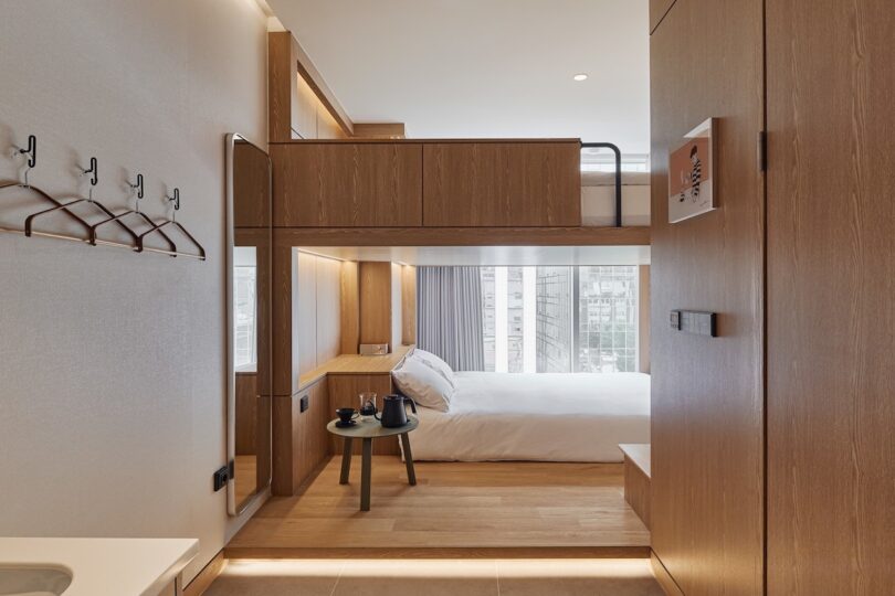 Modern drip and dripping room with wooden furniture, an attic bed above a larger bed, a small table with a kettle to make coffee, wall -mounted hooks and a large window that floods the space with natural light