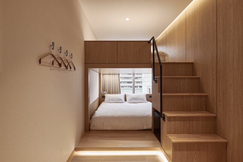 A modern compact bedroom at the Drip & Drop hotel has a double bed under a wooden attic, right stairs and cloth hooks mounted on the left wall. Natural light flows through a window at the distant end