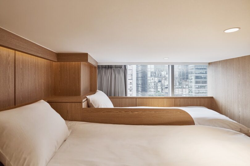Two single beds made with white linen are positioned in a small attic, with wooden panels, at the Drip & Drop hotel, with a large window, overlooking the city buildings in the background.