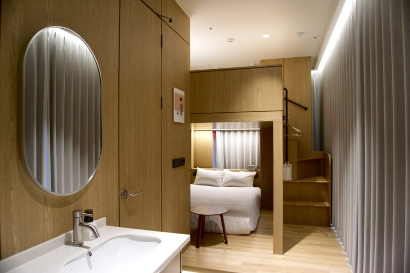Modern hotel room with light wood finishes features a bed, loft area accessible by stairs, a small round table, wall-mounted mirror, and sink in the foreground