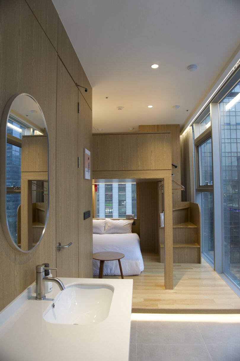 A modern room featuring a built-in wooden loft bed, a white bed below, large windows, and a sink with a round mirror above in the foreground