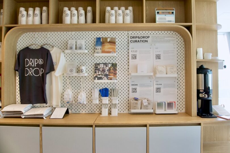 Merchandise and coffee display counter at HOTEL DRIP&DROP with branded gear, paper cups, utensils, brochures, coffee bags, and a coffee dispenser against a light wooden display wall