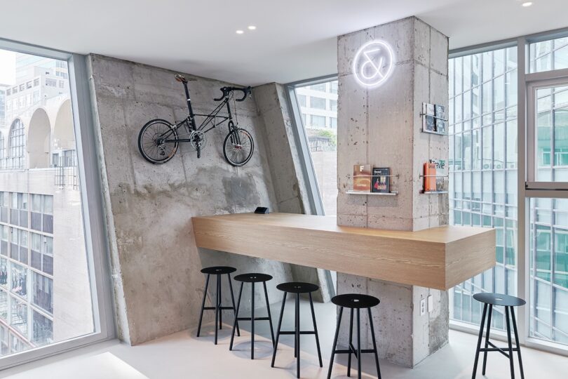 The modern theme of the minimalist cafe at the Drip & Drop hotel with a concrete wall, a wooden countertop and five black chairs, a bicycle mounted on the left wall and a neon sign to the right. Large windows offer views of the city.