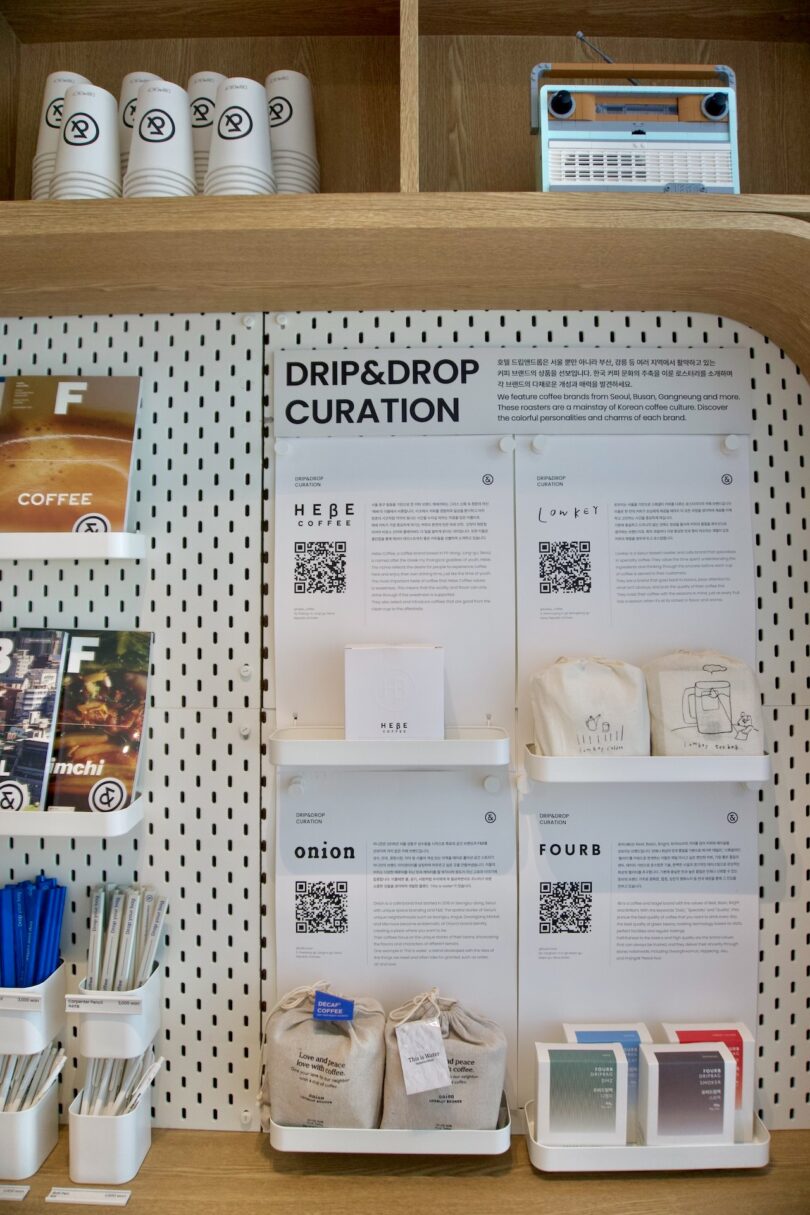 Shelves display coffee bags, tote bags, mugs, and informational cards under a sign labeled "Drip&Drop Curation" at HOTEL DRIP&DROP. Cups and a radio sit on the top shelf