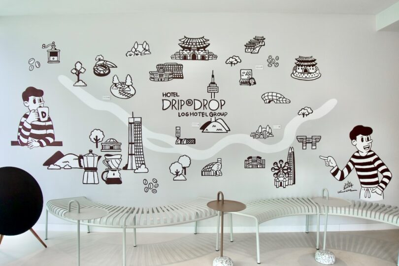 A wall mural in the HOTEL DRIP&DROP lobby features black and white illustrations of buildings, landmarks, and cartoon characters above a row of modern benches