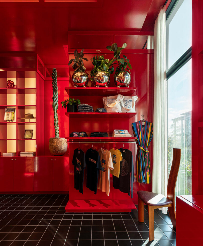 A bright red retail space with hanging clothing, potted plants on shelves, a striped robe, a wooden chair, large windows, and black tiled flooring.