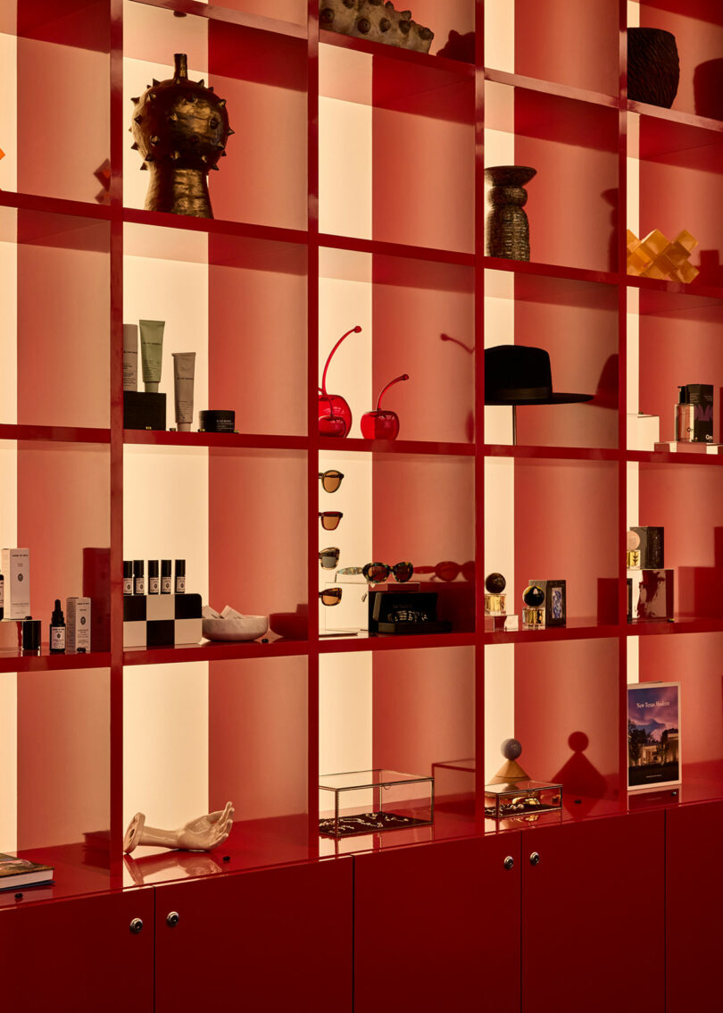 Red shelving unit with various decorative objects, skincare products, sunglasses, hats, and small art pieces displayed in separate cubby-like sections.