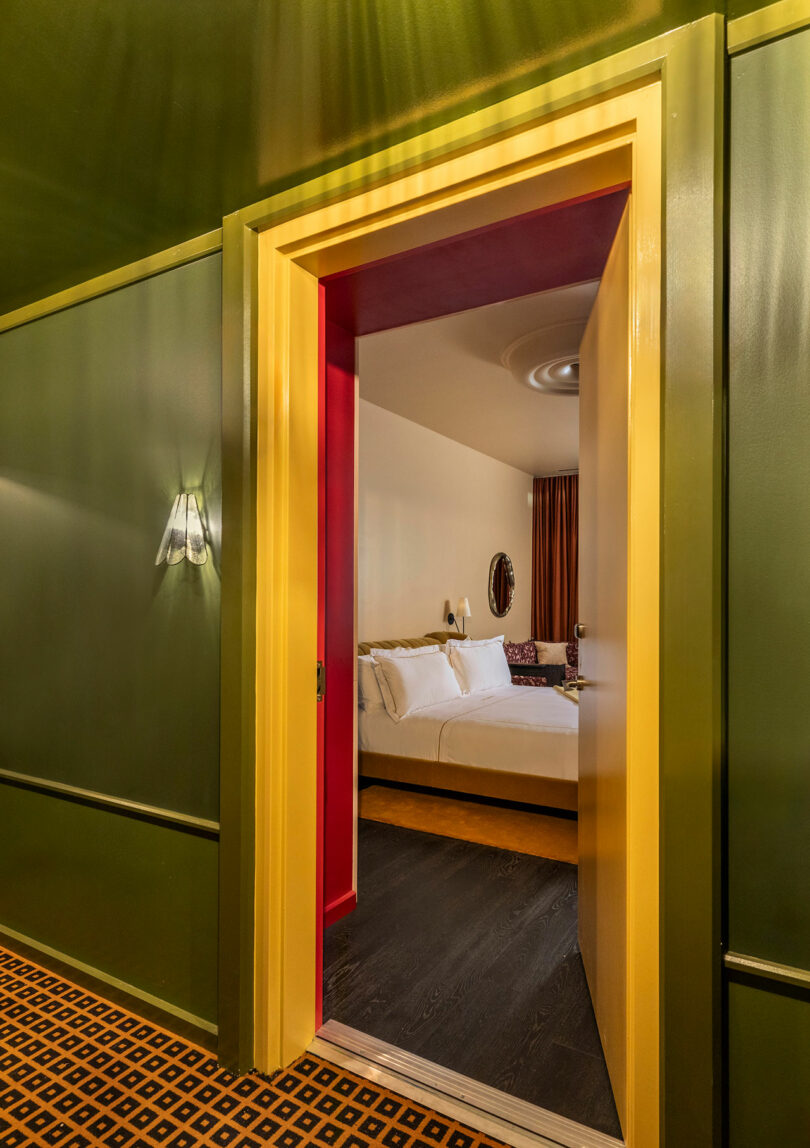 A doorway with yellow trim opens from a green-walled hallway into a bedroom with a double bed, white linens, and warm lighting.