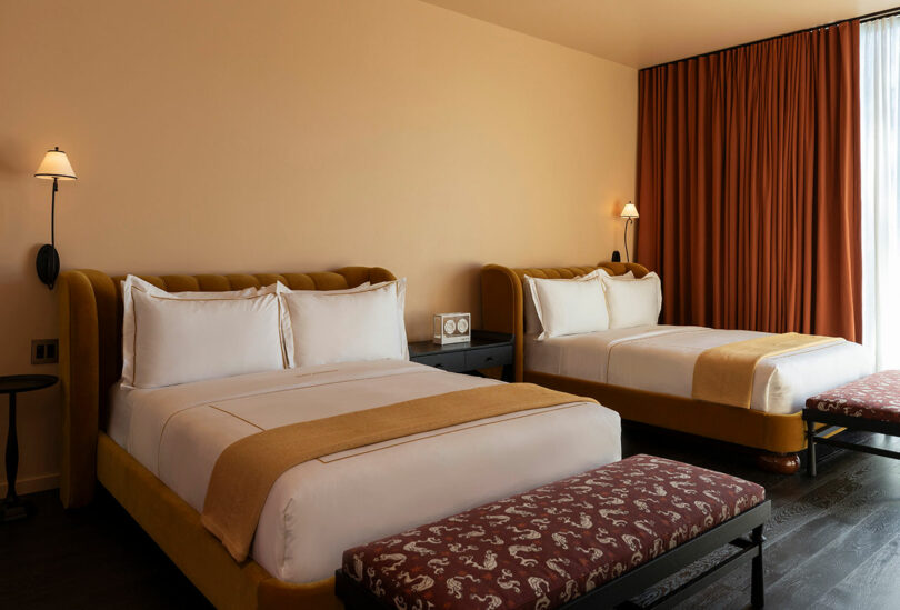 A hotel room with two double beds, white bedding, mustard-colored throws, wall lamps, a nightstand, brown curtains, and patterned benches at the foot of each bed.
