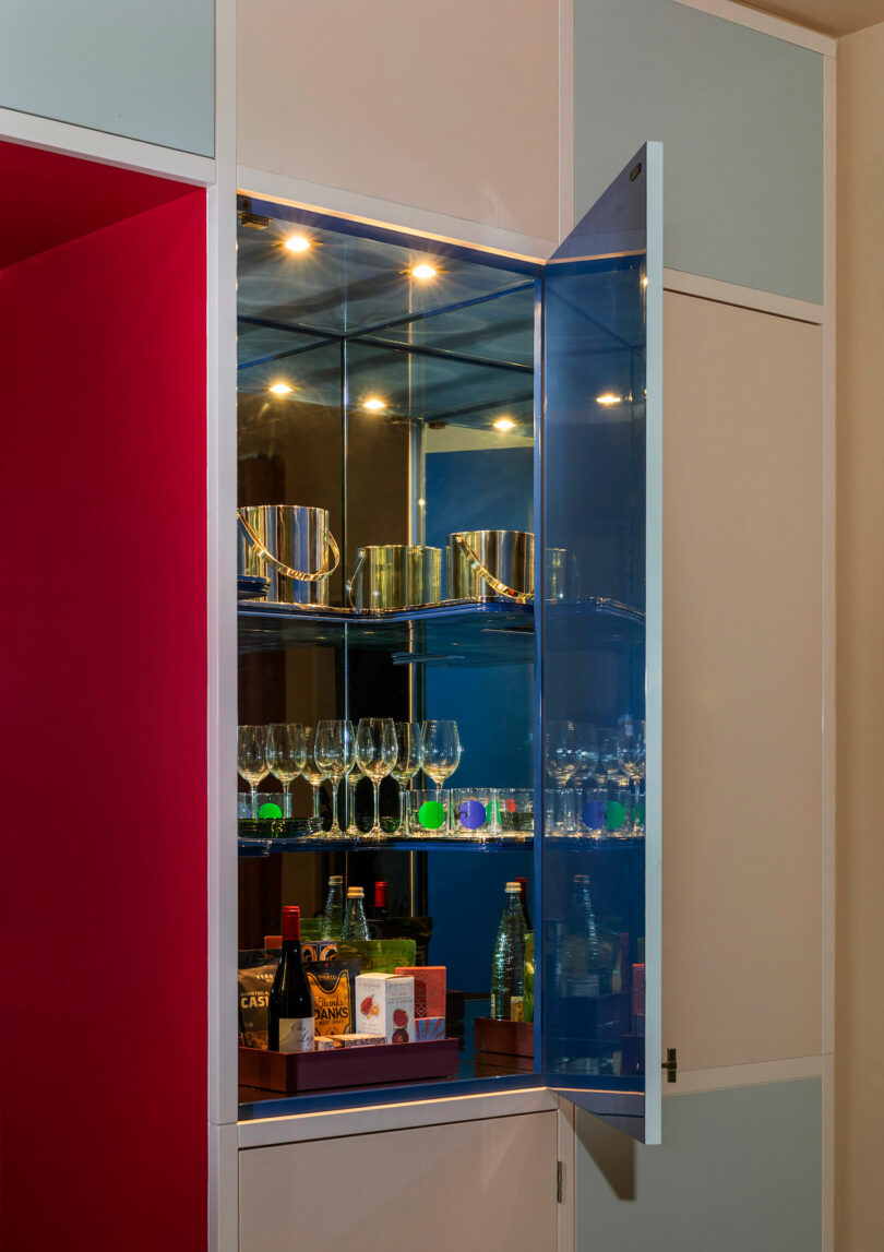 A cabinet with a blue-tinted glass door holds wine glasses, bottles, snacks, and metal ice buckets, with interior lighting illuminating the items.