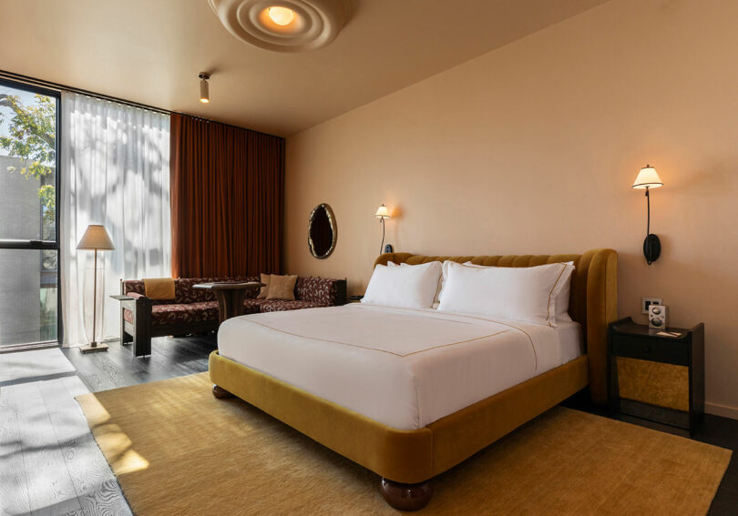 Modern hotel room with a large bed, mustard-yellow headboard and rug, seating area by a window with sheer and brown curtains, and wall-mounted lamps.