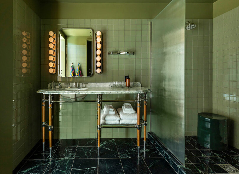 Bathroom with green tiled walls, marble countertop with double sinks, two mirrors with light bulbs, towels on a rack, and a separate shower area with a rain showerhead.