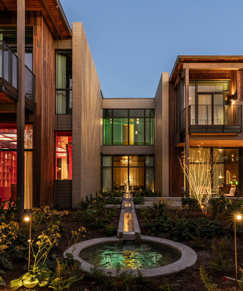 Modern courtyard at dusk with a central water feature, landscaped garden, and two-story building featuring large windows and wooden accents.
