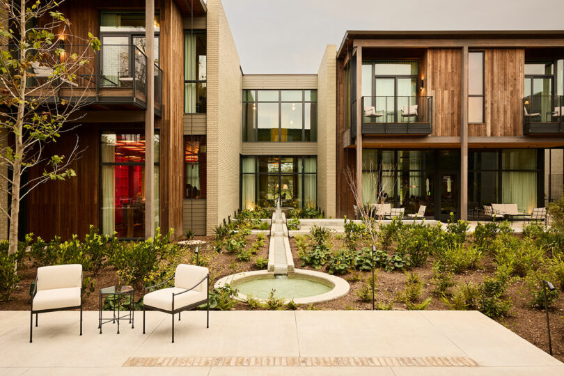 A modern courtyard features landscaped greenery, a small fountain, a water channel, and two cushioned chairs with a side table on a paved area in the foreground.