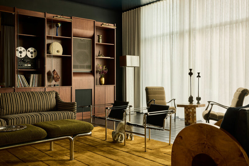 A mid-century modern living room with striped green sofa, glass coffee table, minimalist armchairs, wooden shelving, and floor-to-ceiling sheer curtains.