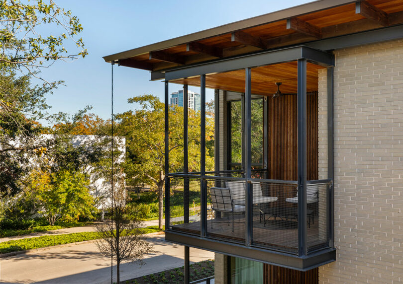 A modern house features a screened balcony with a table and chairs, surrounded by trees and overlooking a quiet residential street.