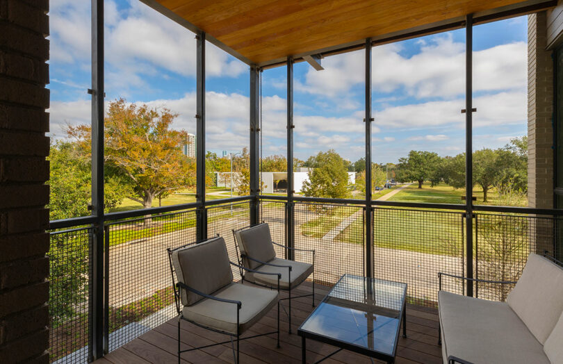 A screened balcony with cushioned chairs and a glass coffee table overlooks a green lawn, trees, and a modern white building under a partly cloudy sky.