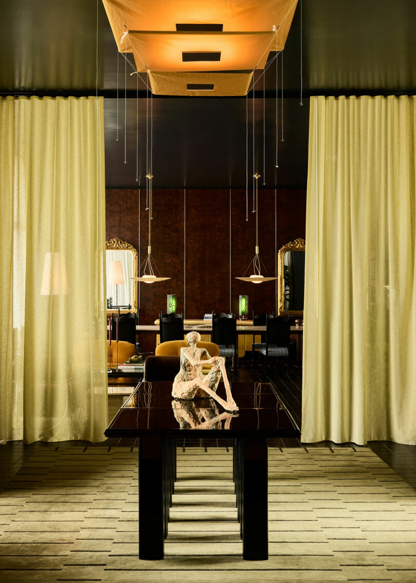 A modern dining room features a black table with a beige abstract sculpture, flanked by sheer curtains and gold accents, under a geometric ceiling light fixture.