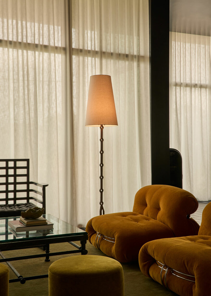 A living room with mustard-colored lounge chairs, a matching ottoman, a glass coffee table with books, a tall floor lamp, and sheer curtains covering large windows.