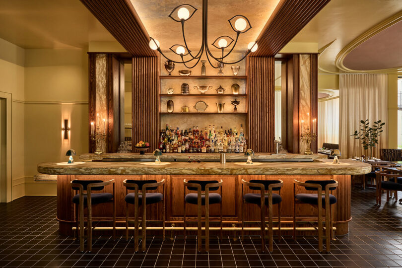 A modern bar with marble countertop, five high-backed stools, shelves displaying bottles and glassware, decorative lighting, and elegant wood and brass accents.